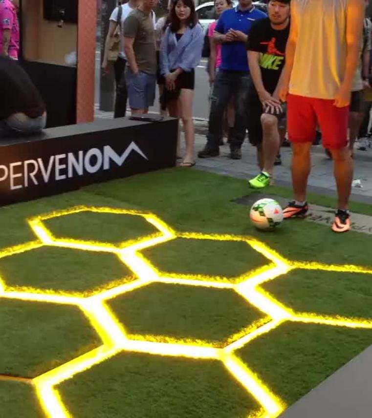 A player dribbling a soccer ball through illuminated yellow hexagon light grids on artificial turf in a Football Light Grid Challenge.