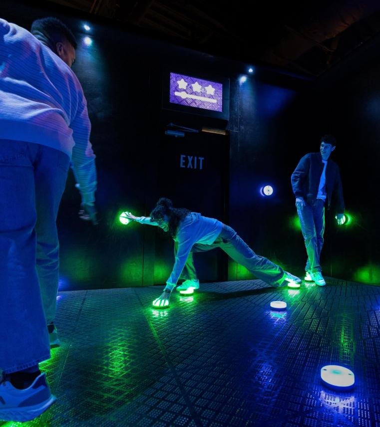 Players stretching to press glowing green LED buttons on the wall and floor simultaneously in an Octopus challenge room.
