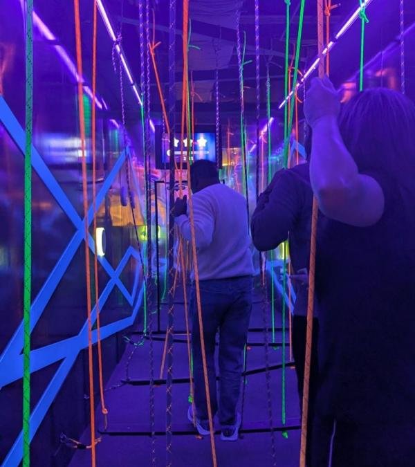 Players navigating through hanging UV ropes and balancing wooden steps inside the Rope Balance Challenge Swing Crossing game room.